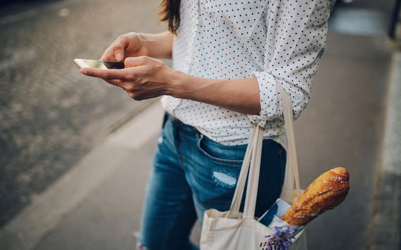 woman walks down the street and texts with a bag of groceries on her arm