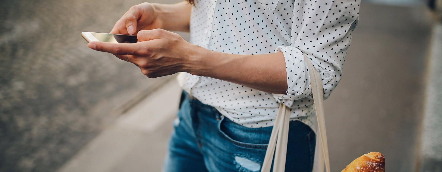 woman walks down the street and texts with a bag of groceries on her arm